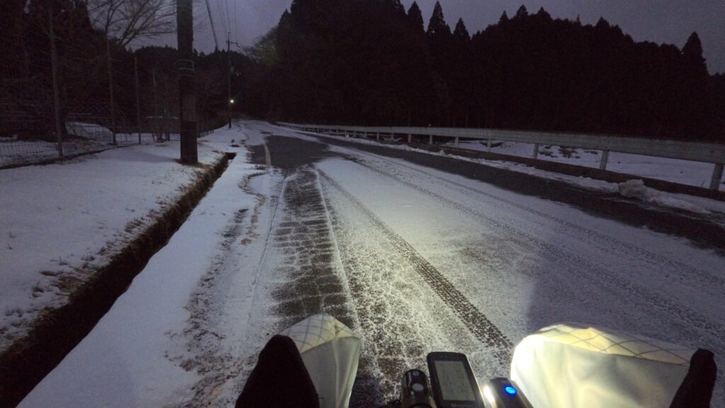 雪が降り始めた夜明け前の道の写真。自転車のハンドルとライトが映り、雪に覆われた車道が続いている。